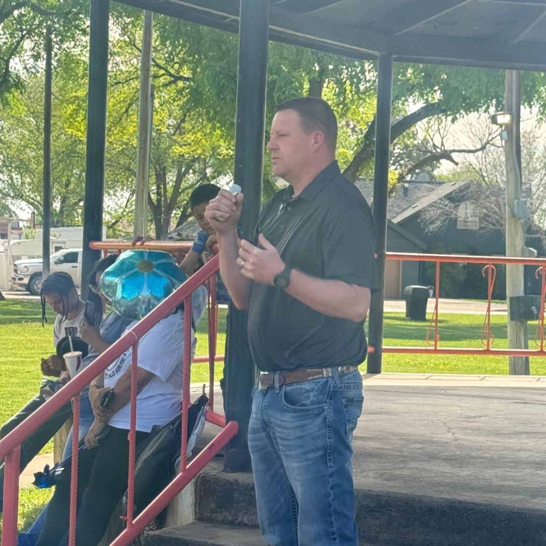 Man speaking outdoors beside a red railing, with people seated in the background under a shaded pavilion