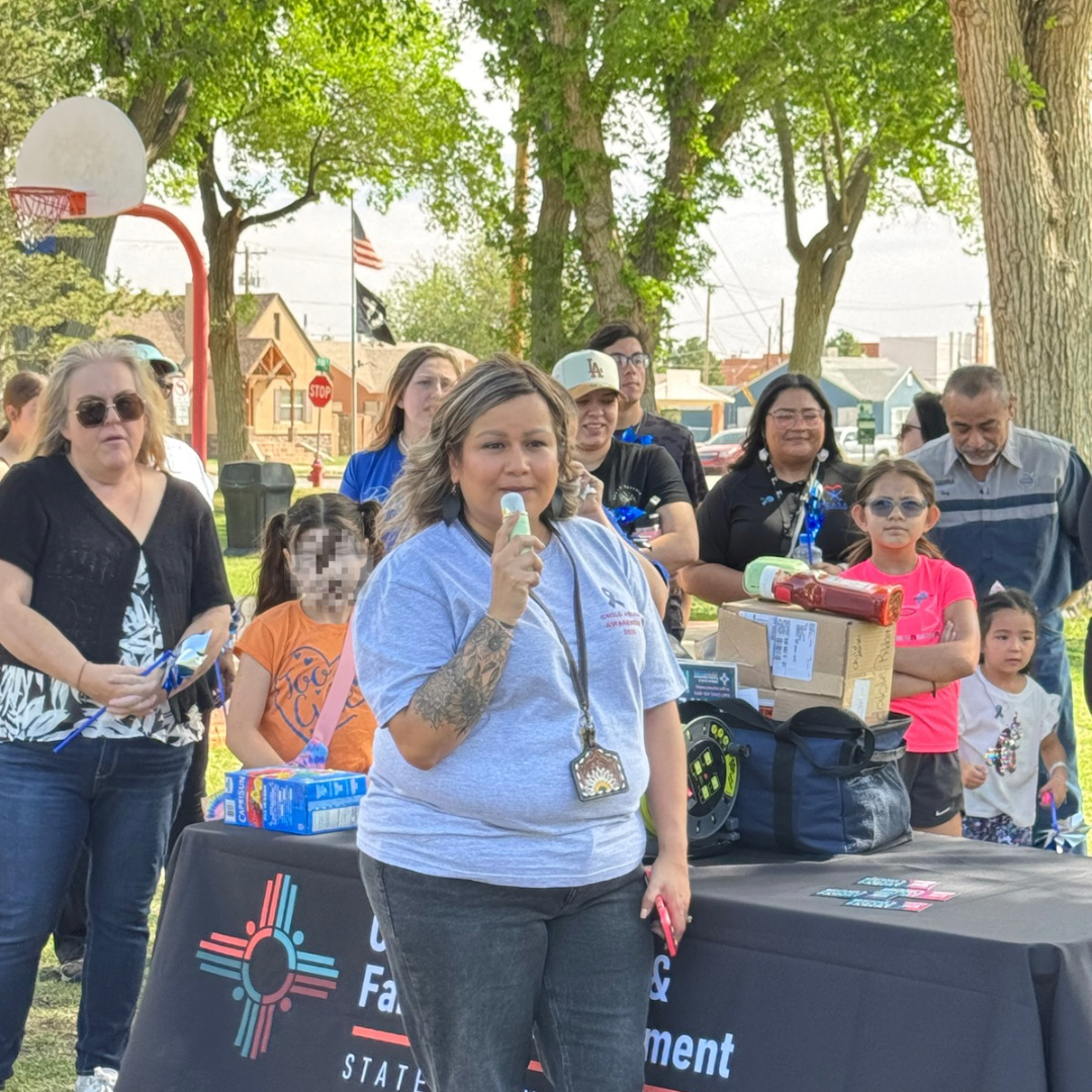 Woman speaking into a microphone at an outdoor community event beside a table with volunteers and supplies