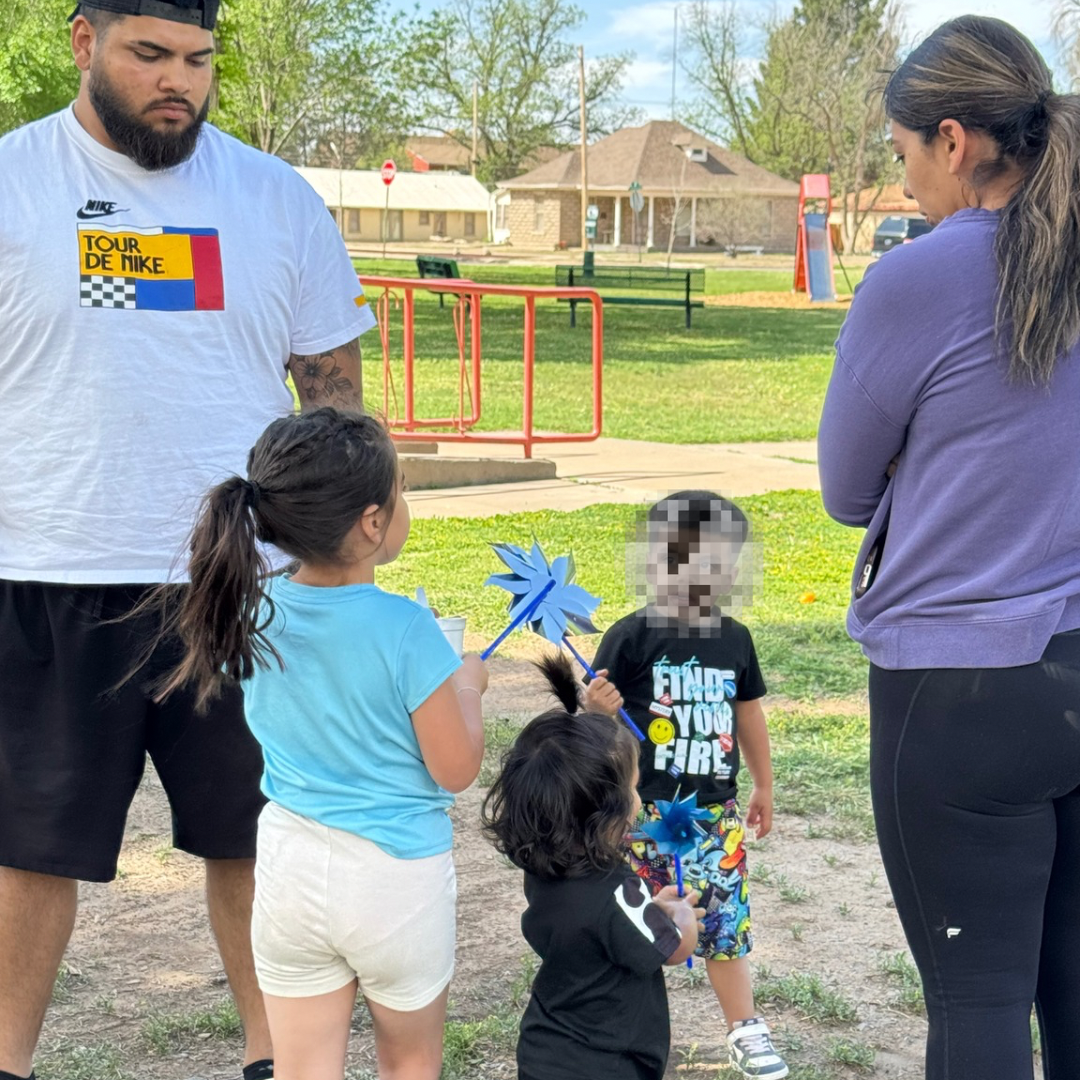 Adult and three children stand in a park near a red fence, with one child holding a blue pinwheel.