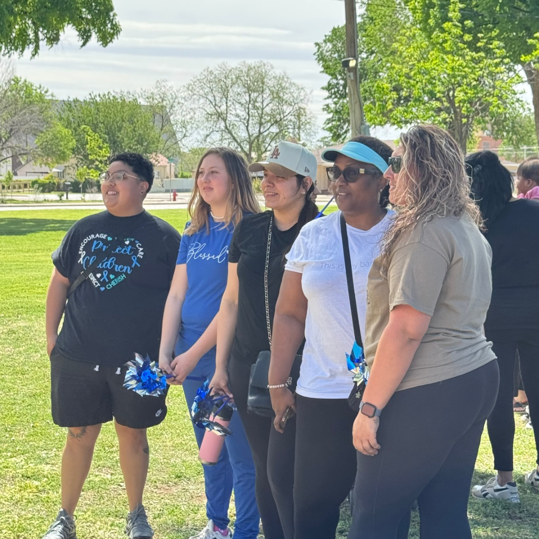 Five people standing outdoors in a park, holding blue pom-poms, posing together in the sun.