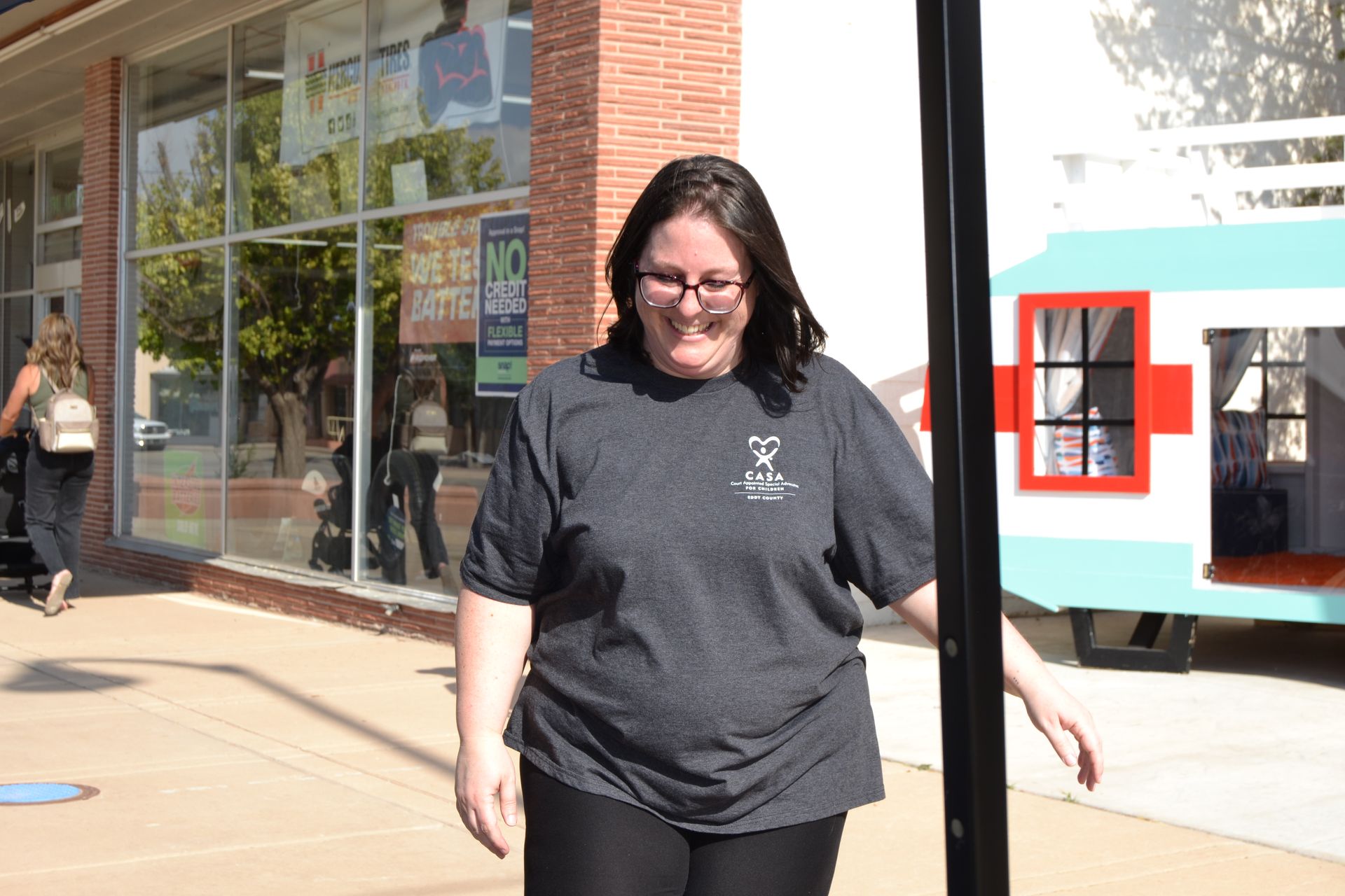 Woman walking on a sunny sidewalk beside a brick building, wearing a dark T-shirt and glasses.