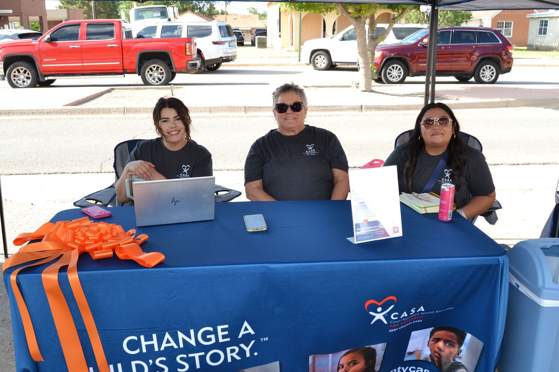 Three people behind a blue tablecloth at a community outreach booth outdoors.
