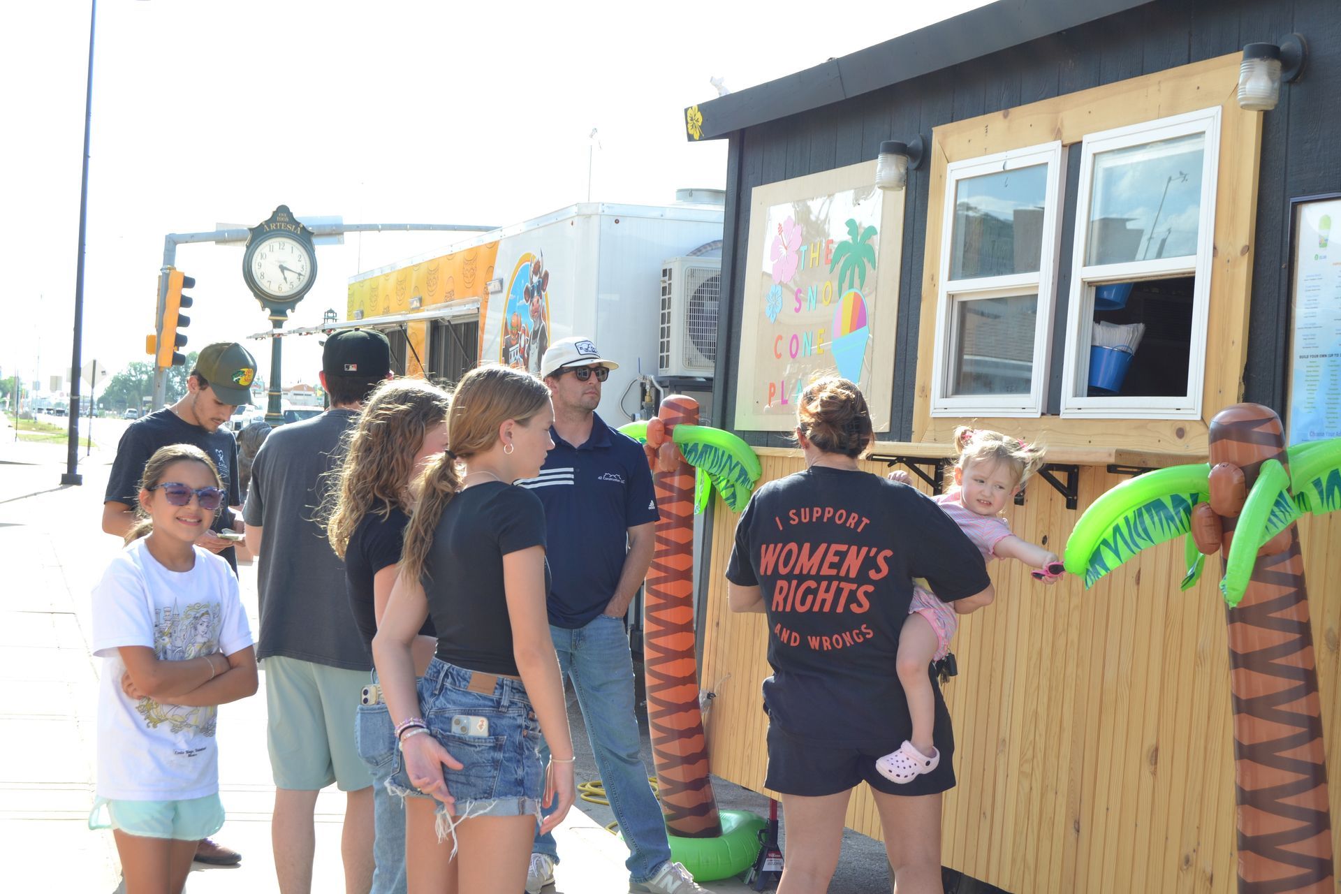 People line up outside a small kiosk, with a wooden counter, colorful posters, and a green statue-like figure.