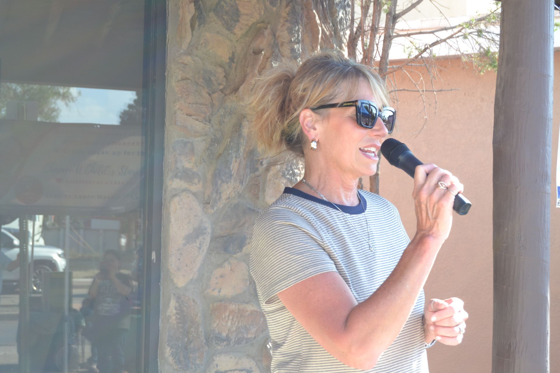 Woman in sunglasses speaking into a microphone outdoors beside a stone column