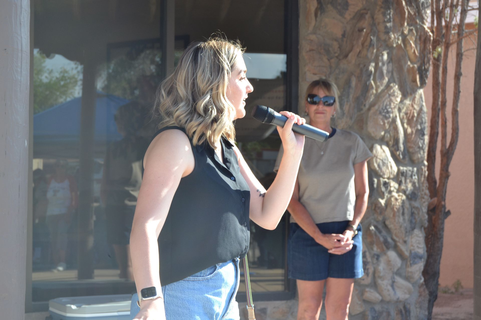Woman singing into a microphone outdoors while another person stands nearby in sunglasses.