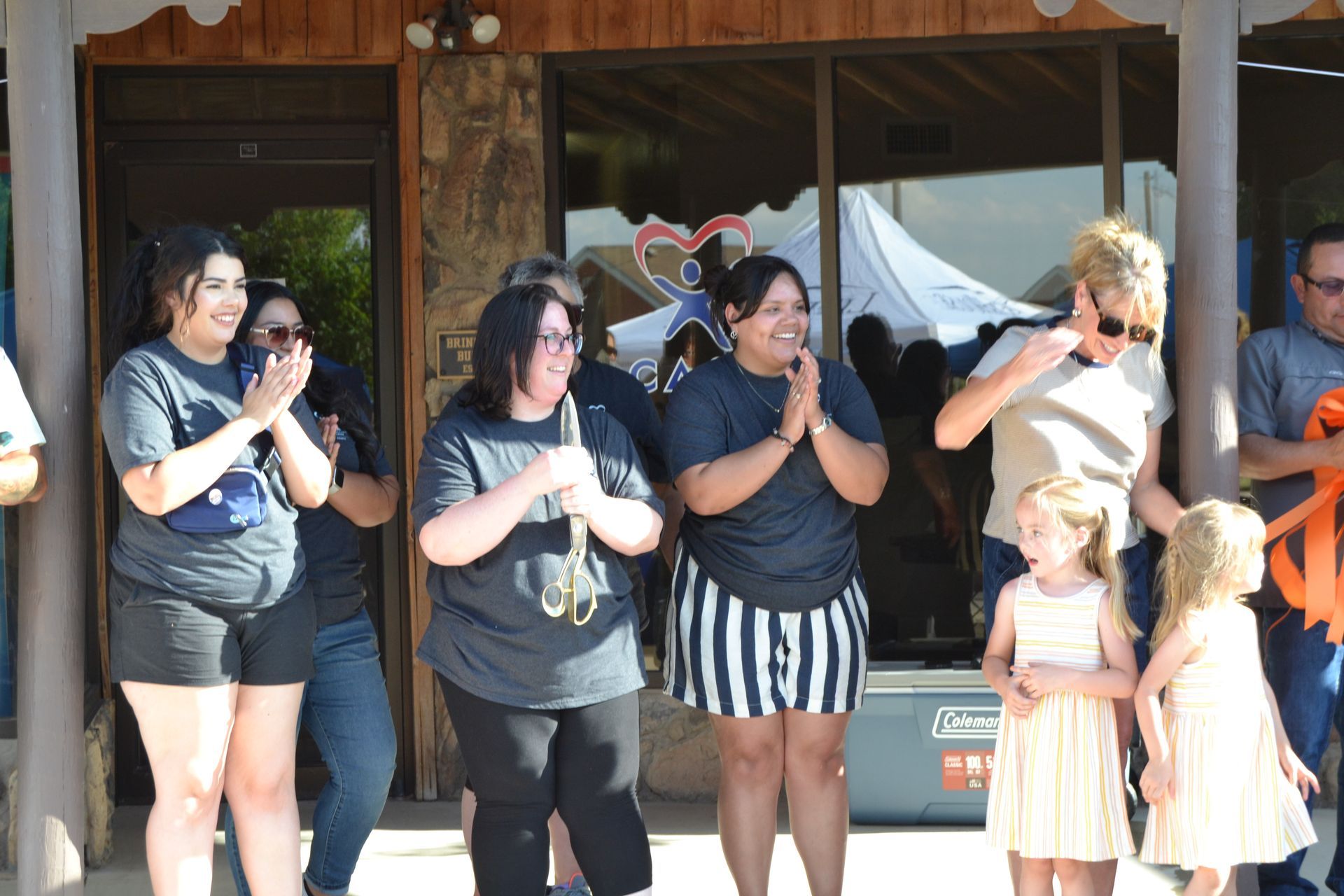 People clapping outside a wooden building, with two children standing nearby.