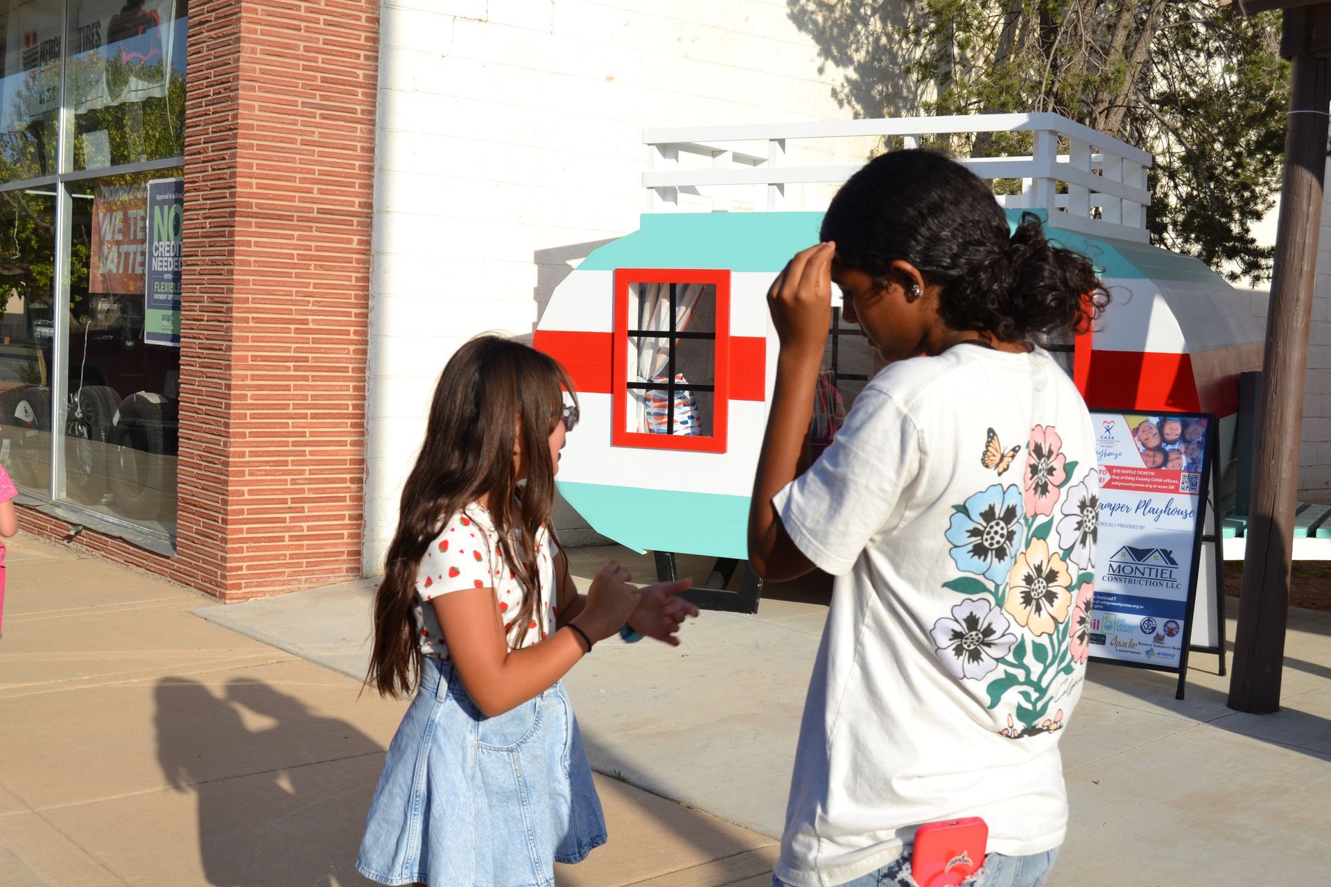 Two children stand outside by a colorful playhouse display on a sunny sidewalk.