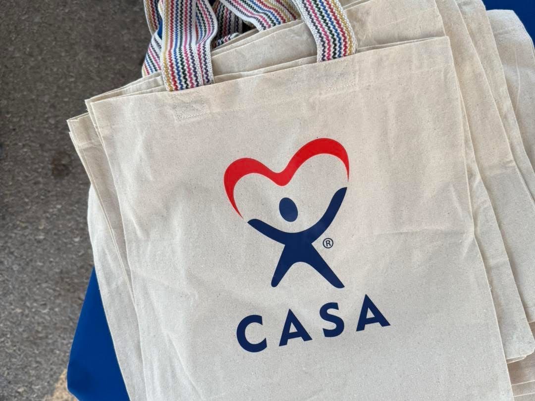 White CASA tote bag with blue and red logo and striped handles on a dark surface