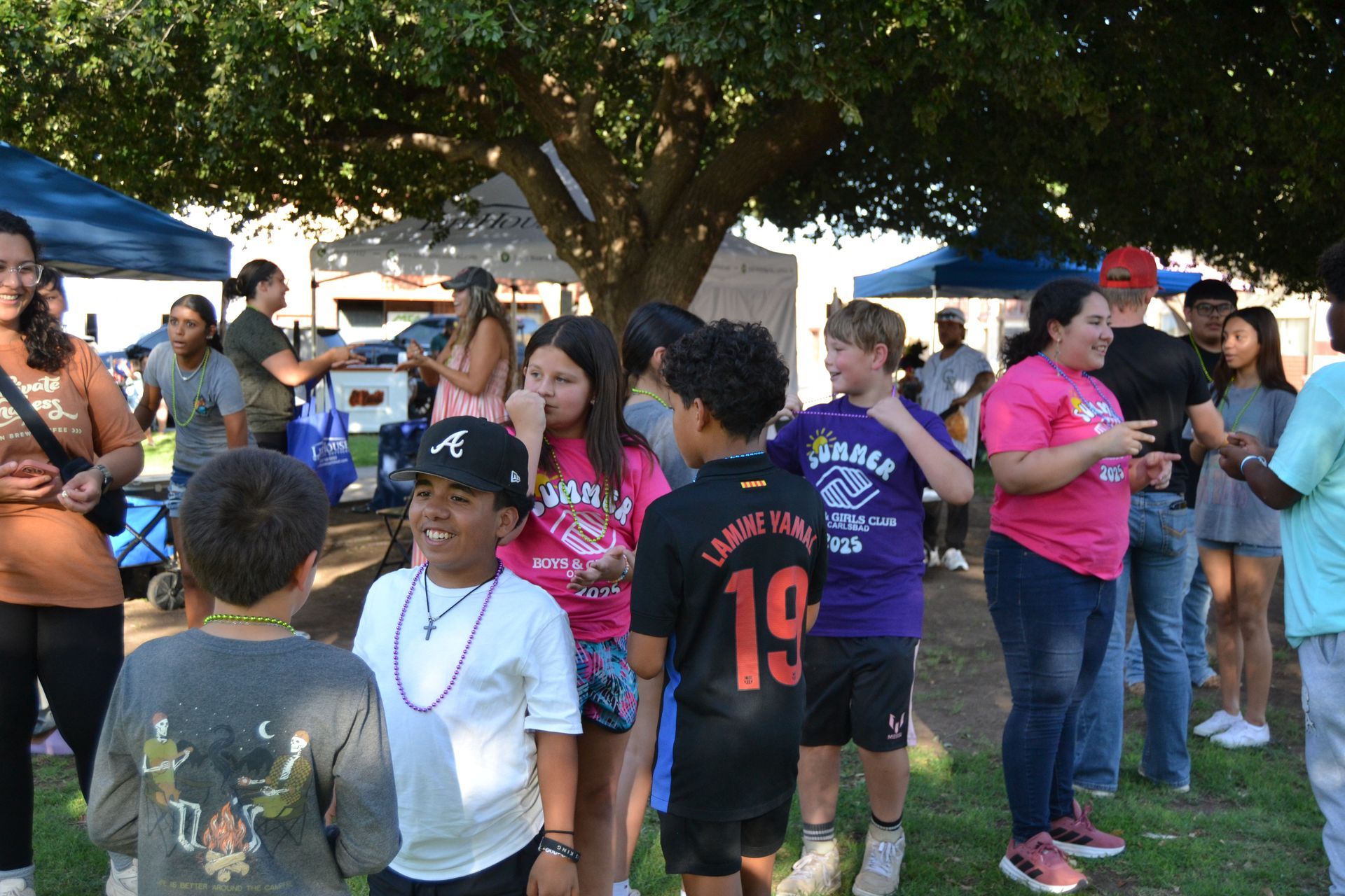 People gathered outdoors under a tree, wearing colorful shirts and chatting at a community event.