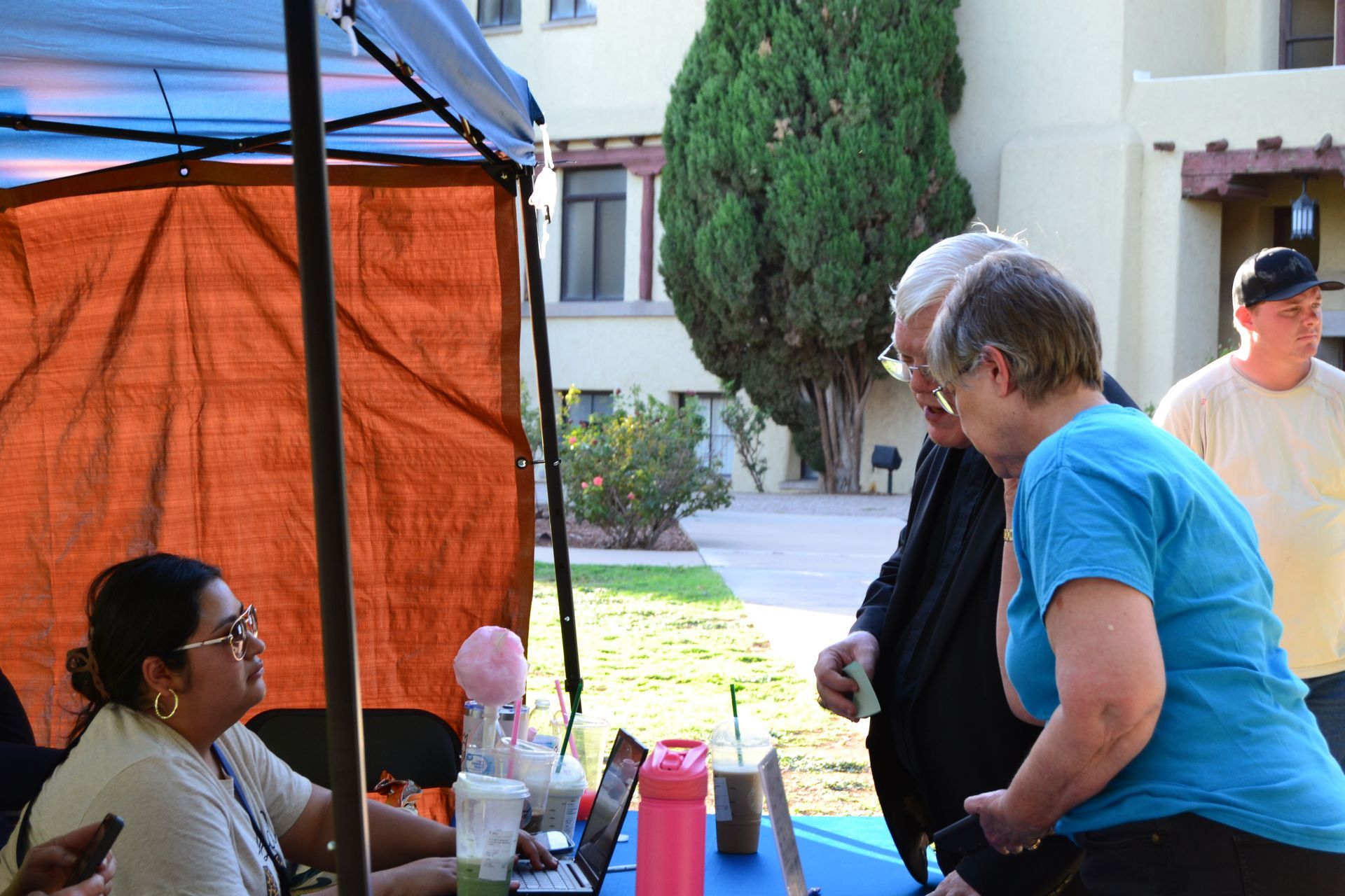 People talking at an outdoor booth under a canopy, with tables, drinks, and a courtyard in the background.
