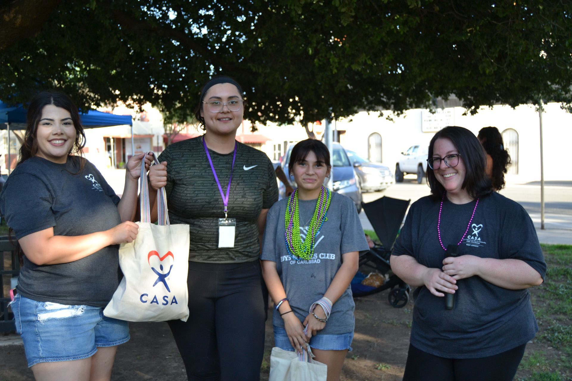Four people stand outdoors holding a CASA tote bag and certificates under a shady tree.