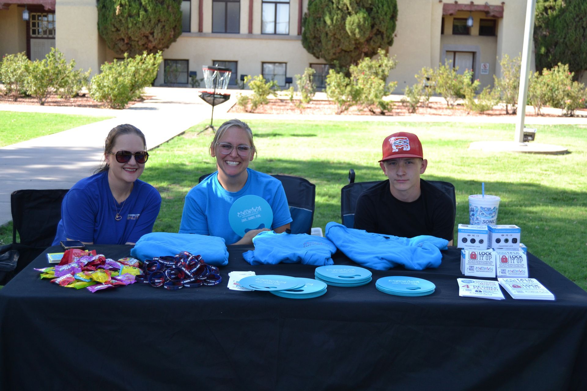 Three people behind a table with blue shirts, snacks, and folded T-shirts at an outdoor event