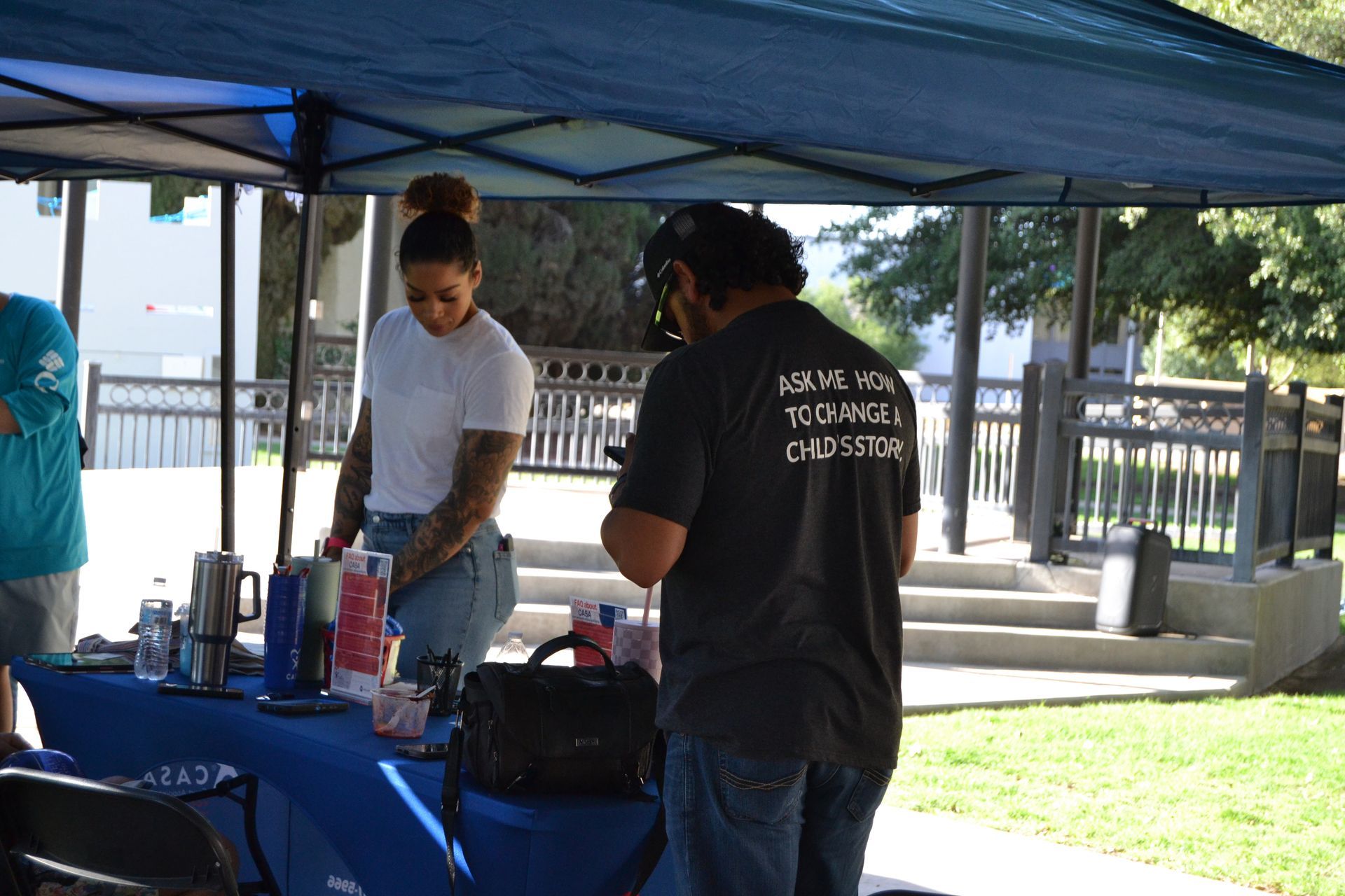People standing at a blue table under a canopy, setting up items outdoors in a park.