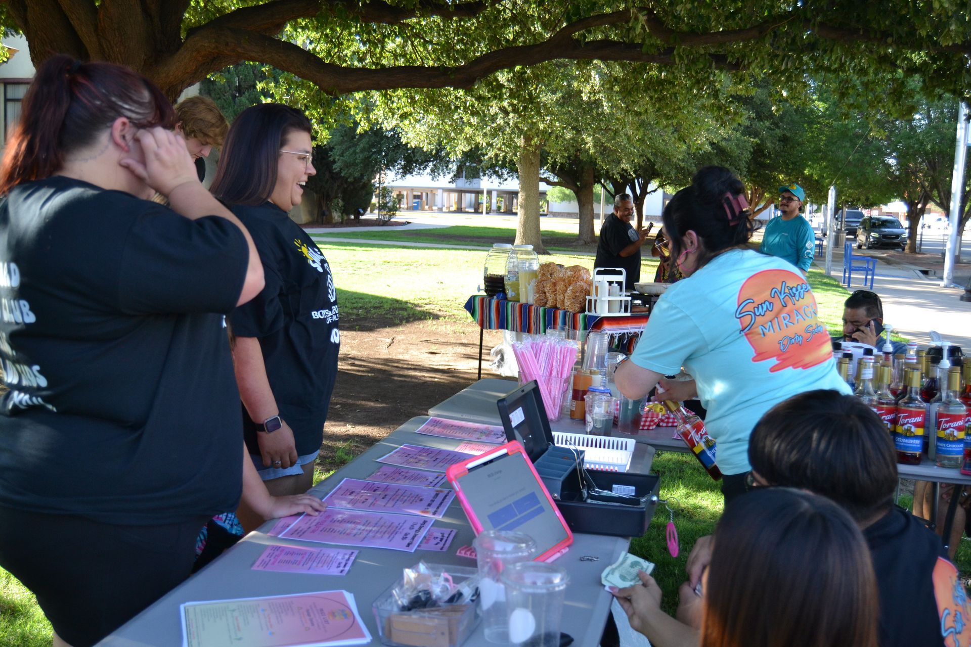 People browsing a vendor table with colorful items at an outdoor market under a large tree