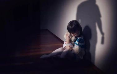Child sitting alone on the floor in a dim hallway, hugging knees, with a shadow on the wall.