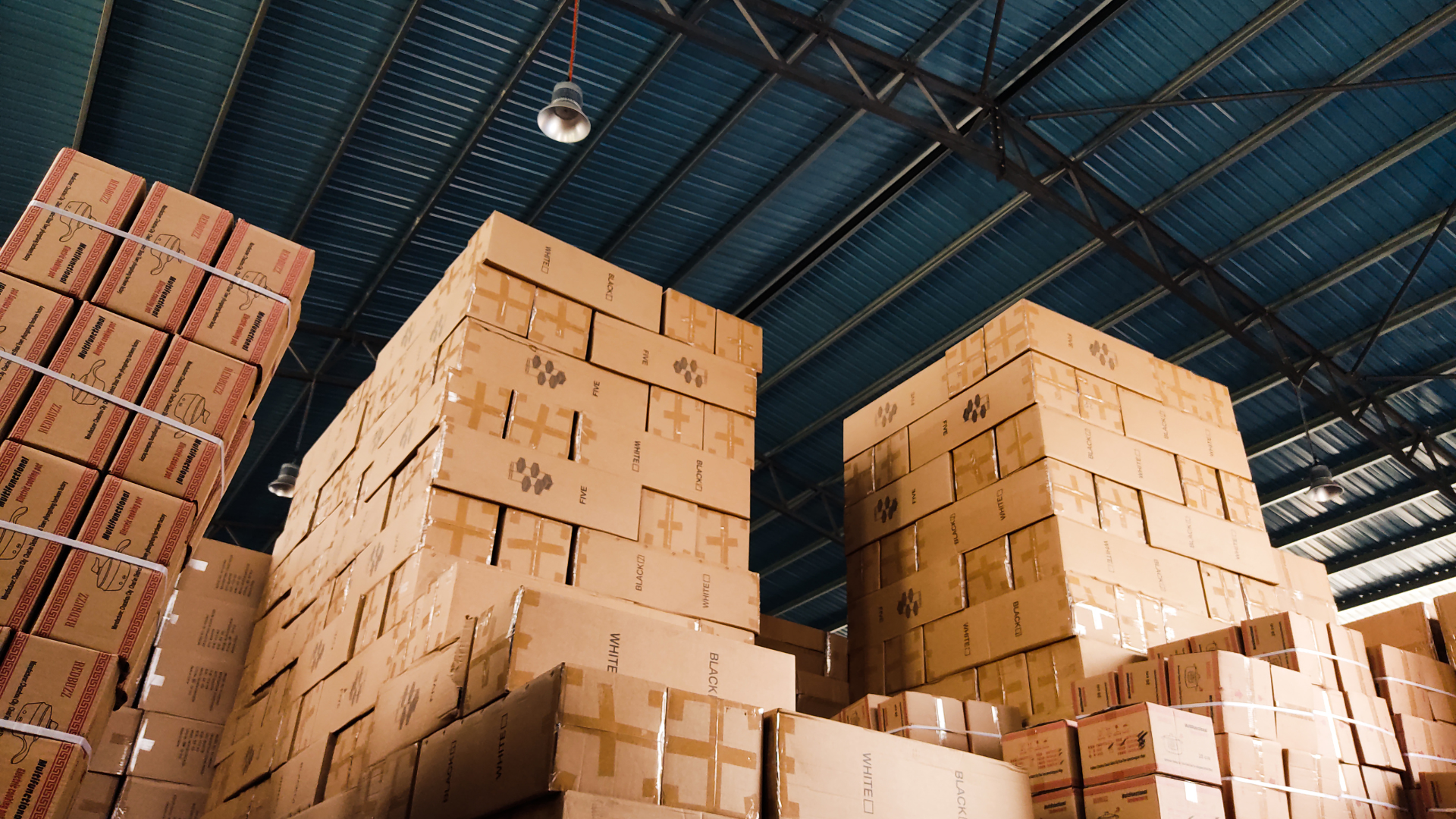 Des cartons empilés en hauteur dans un entrepôt au toit métallique.