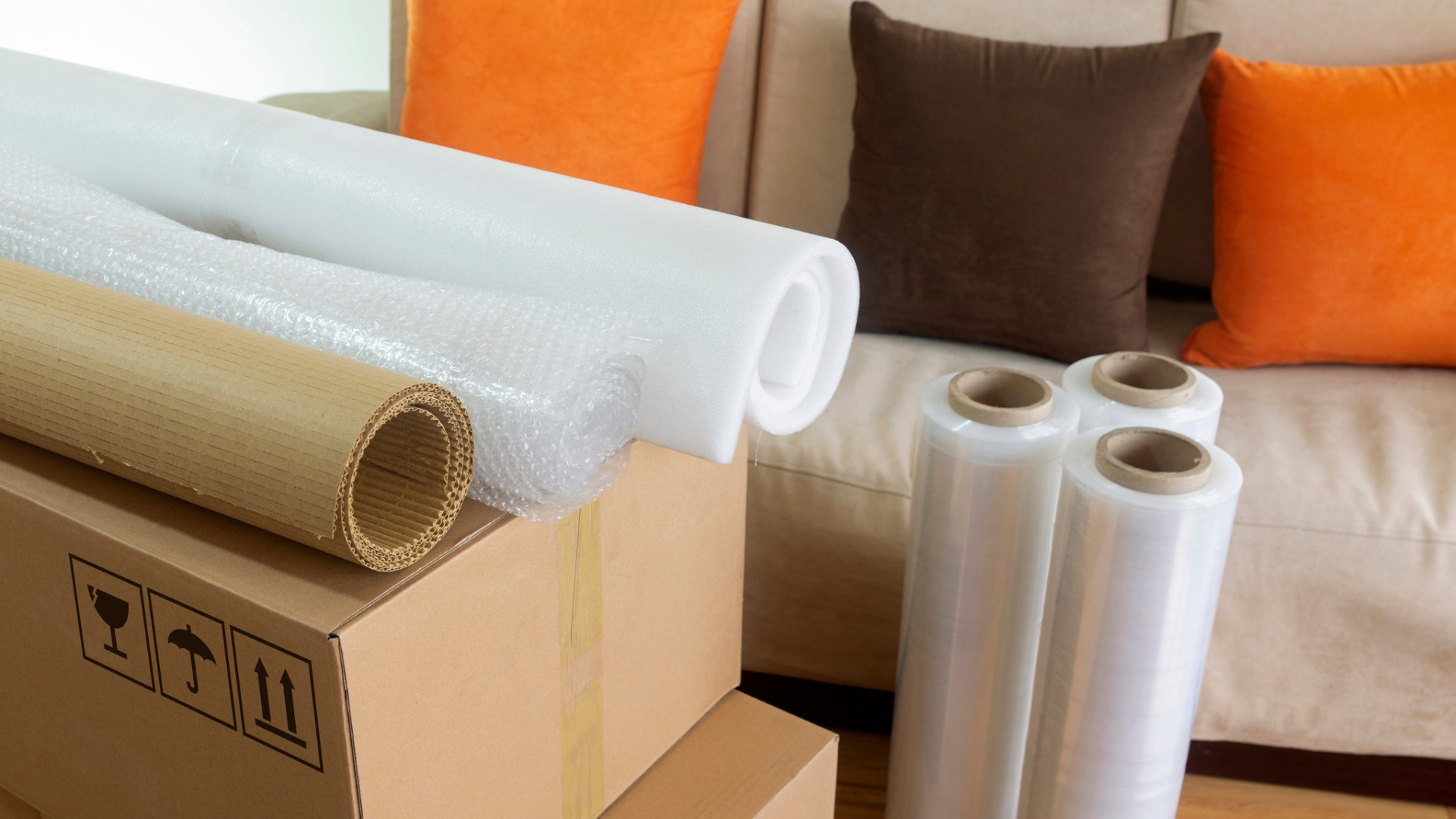 Fournitures de déménagement : cartons, rouleaux de papier bulle, ruban adhésif d’emballage, devant un canapé avec des coussins orange et marron.