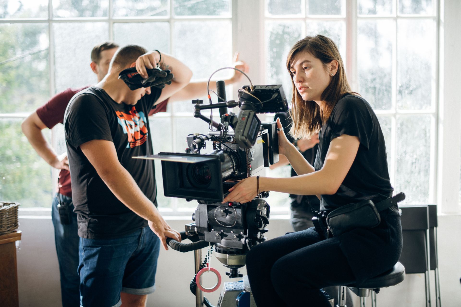 Crew filming with a camera rig in a bright studio, with one person adjusting equipment and another seated nearby.