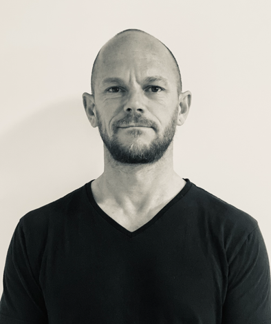 Portrait of a bald man with a beard wearing a black V-neck shirt against a plain light background