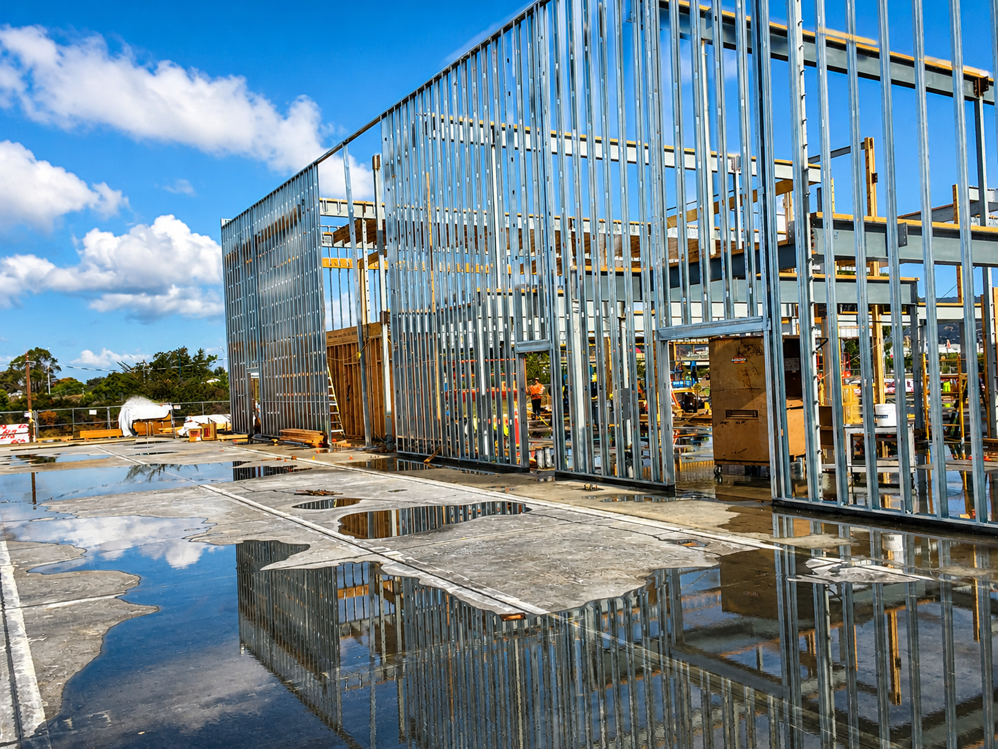 Steel-framed building under construction with puddles reflecting the structure and blue sky