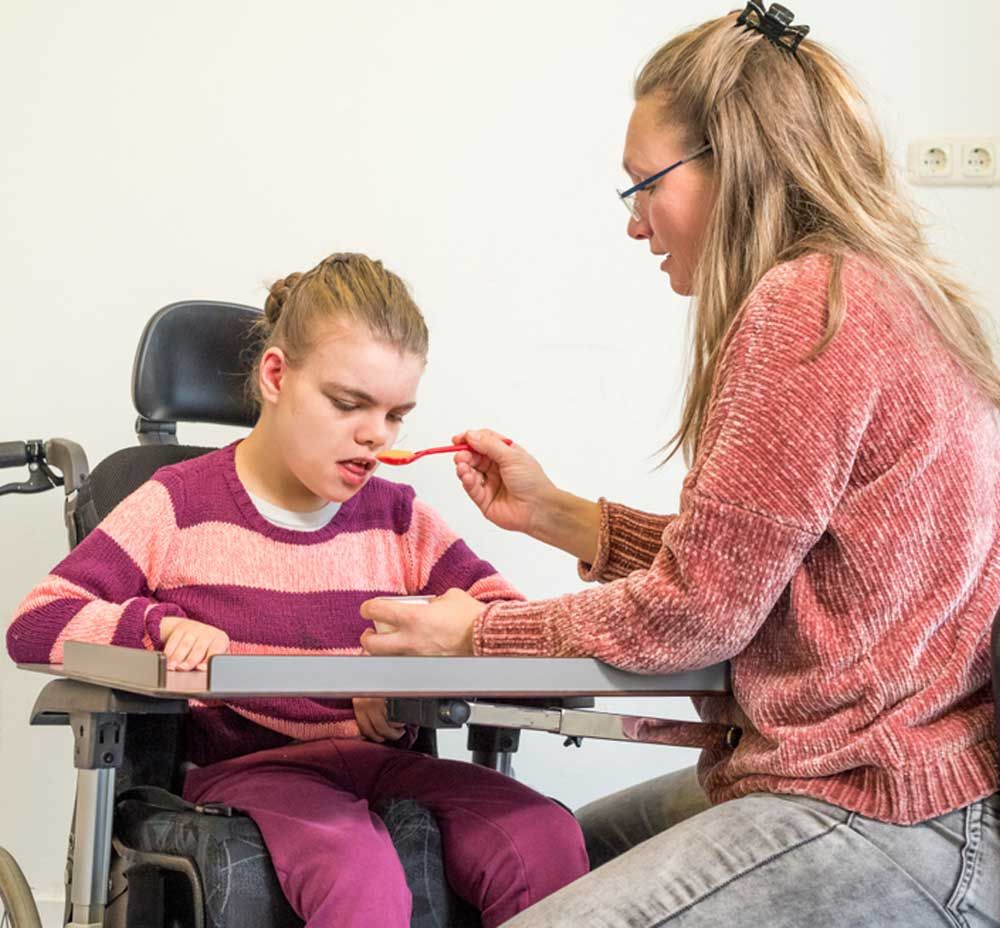A Woman Is Feeding a Child in A Wheelchair with A Spoon — Active Ables Community Services In Inverell, NSW