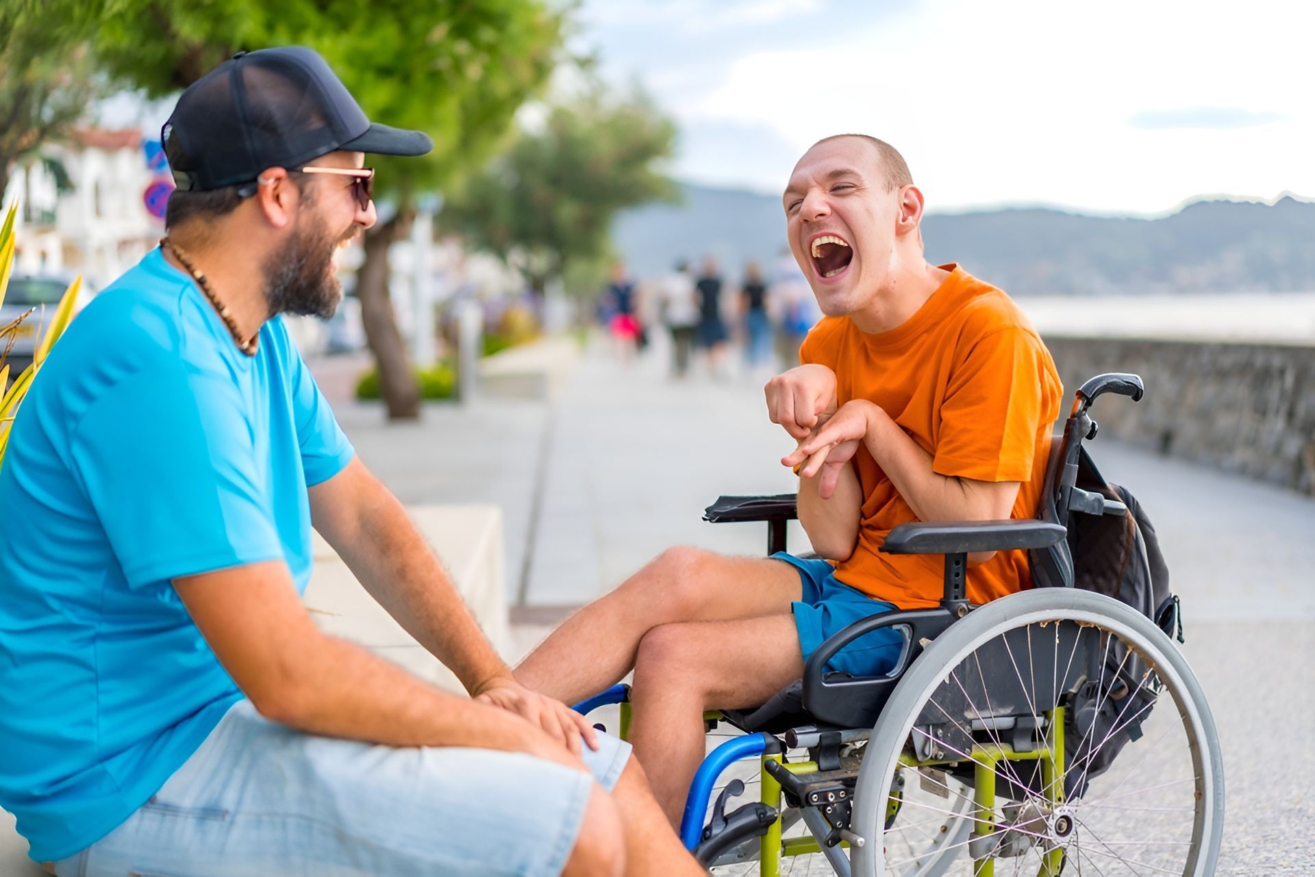 Man in blue shirt talking with laughing man in wheelchair on a waterfront promenade