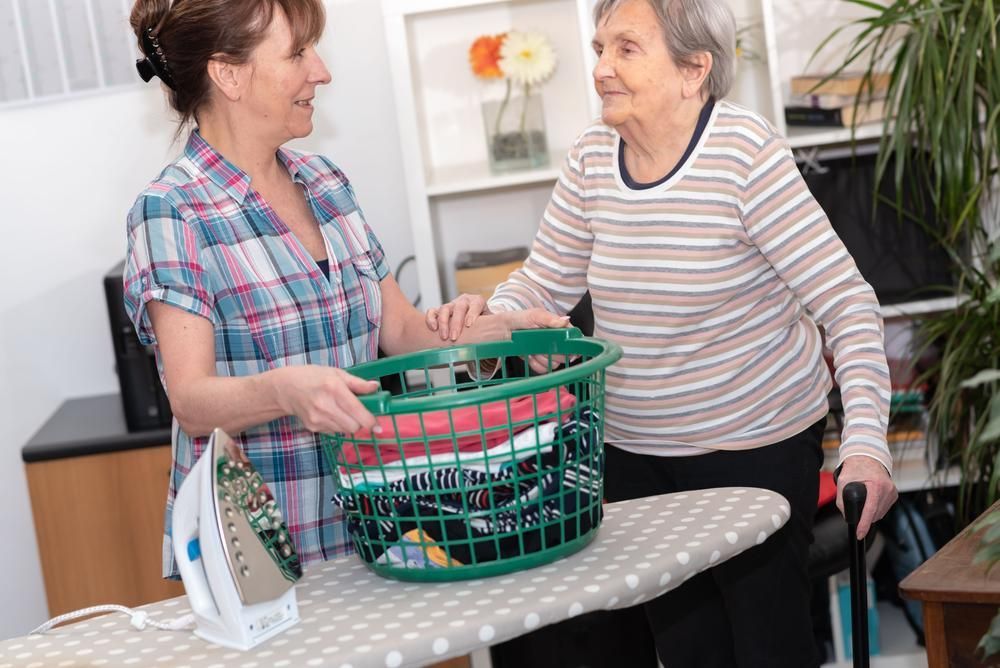 A Woman Is Helping An Elderly Woman Iron Clothes On An Ironing Board — Active Ables Community Services In Tamworth, NSW
