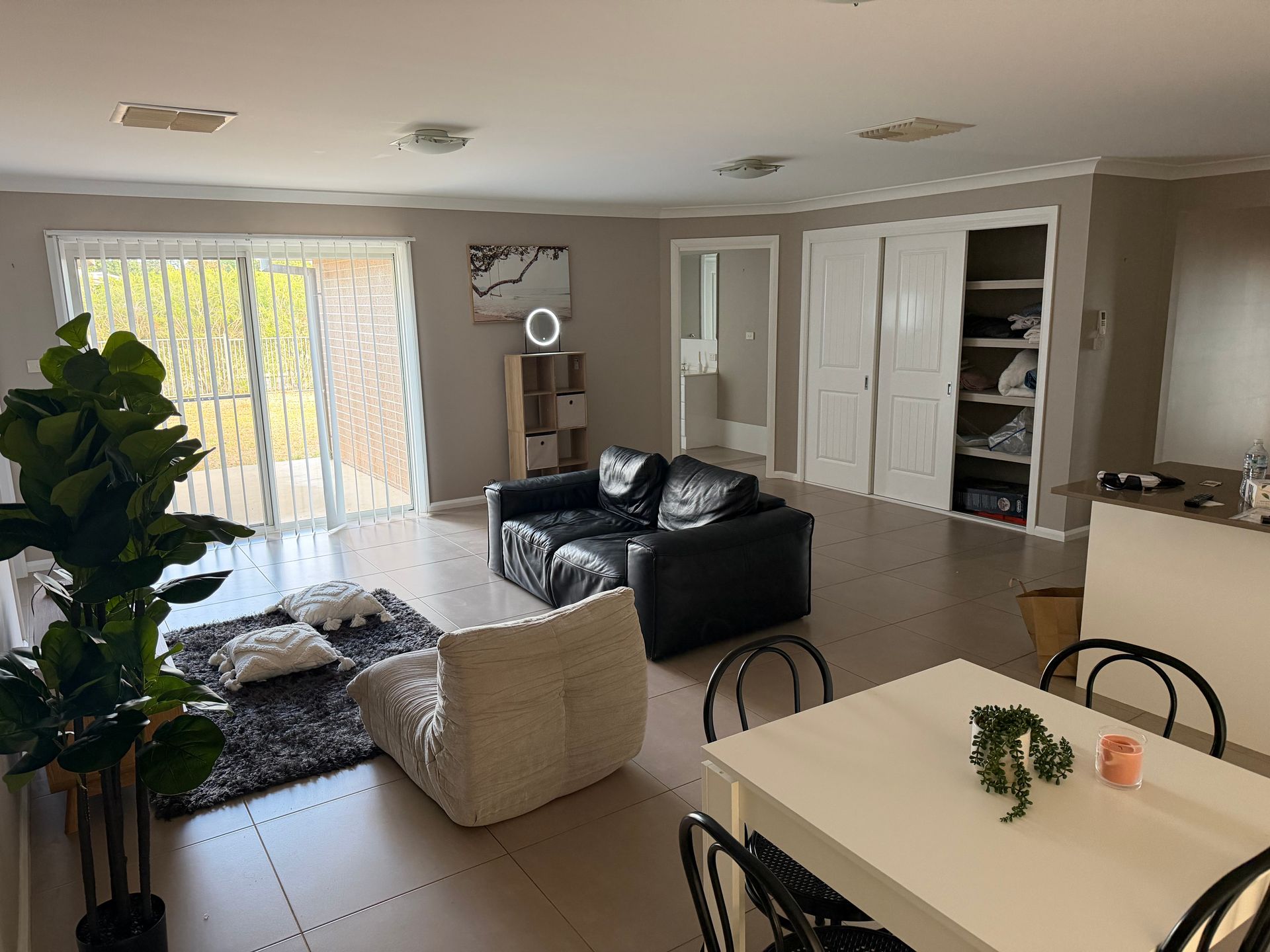 Bright living room with black sofas, a white chair, dining table, sliding glass doors, and open shelving.