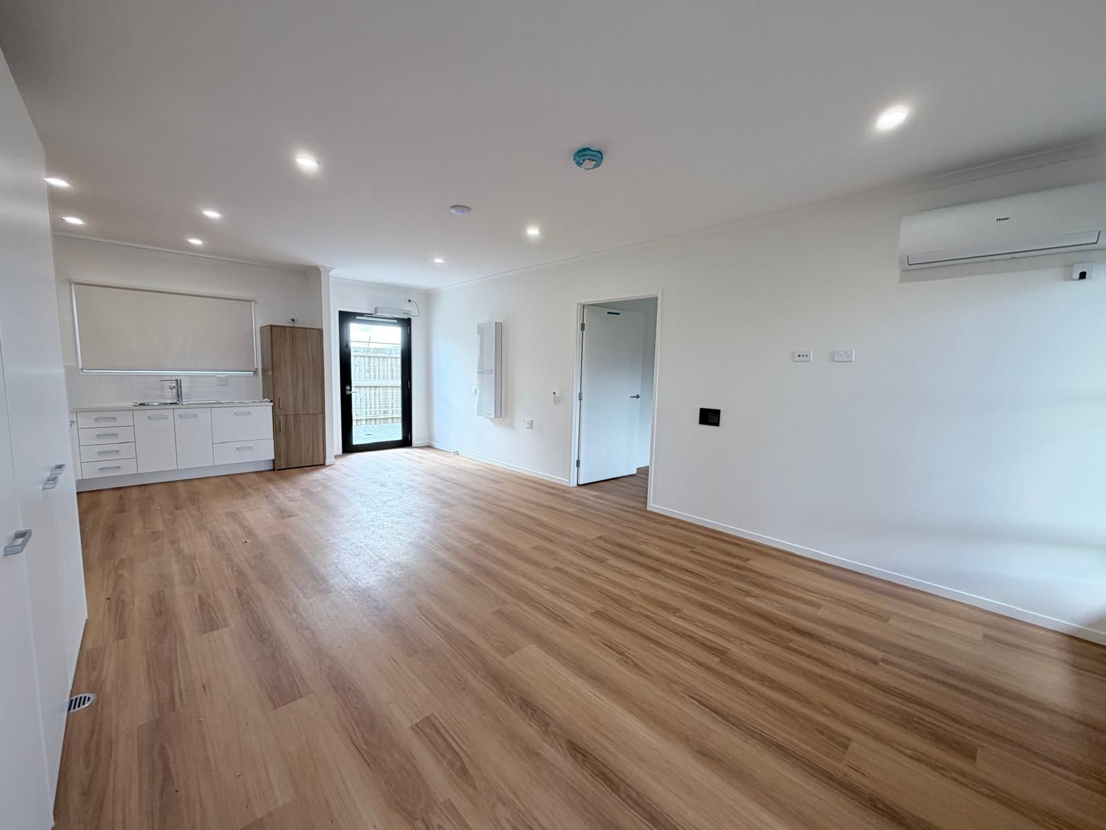 Empty modern apartment living area with wood floors, white walls, kitchen, and wall-mounted AC unit