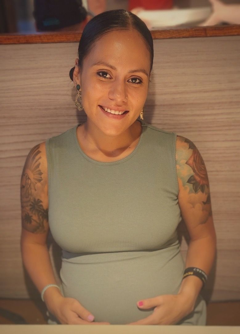 Smiling person in a green sleeveless top, seated indoors with tattooed arms and hands resting on a pregnant belly