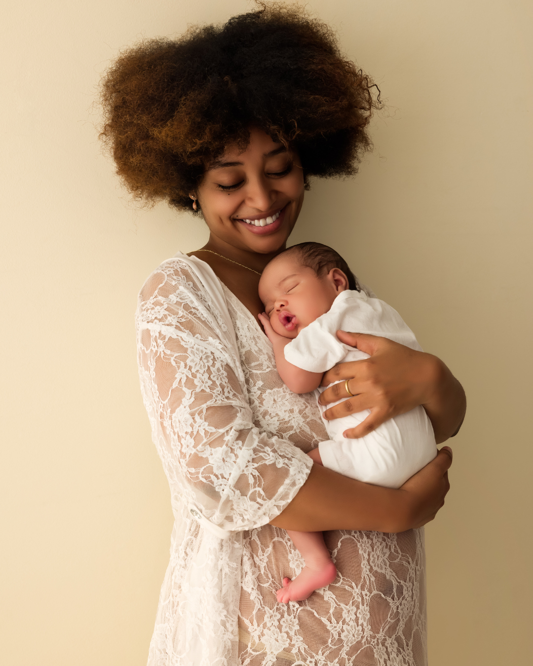 Smiling adult holding a sleeping baby in a white lace dress against a beige background