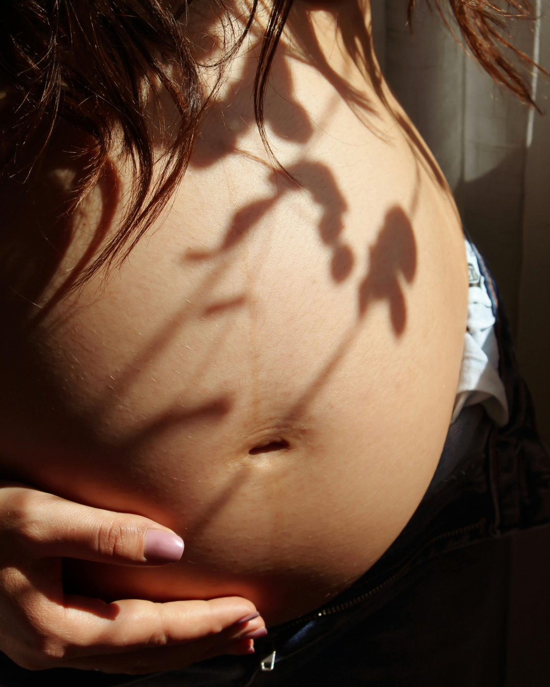 Close-up of a bare pregnant belly with hands resting below and leaf shadows across the skin