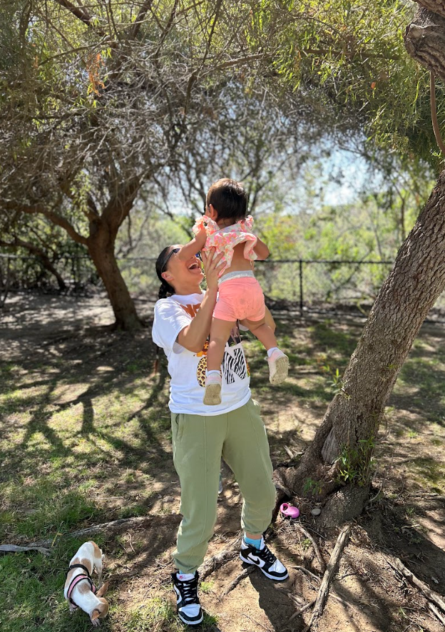 Adult lifting child under trees in a sunny park, with a small dog nearby.