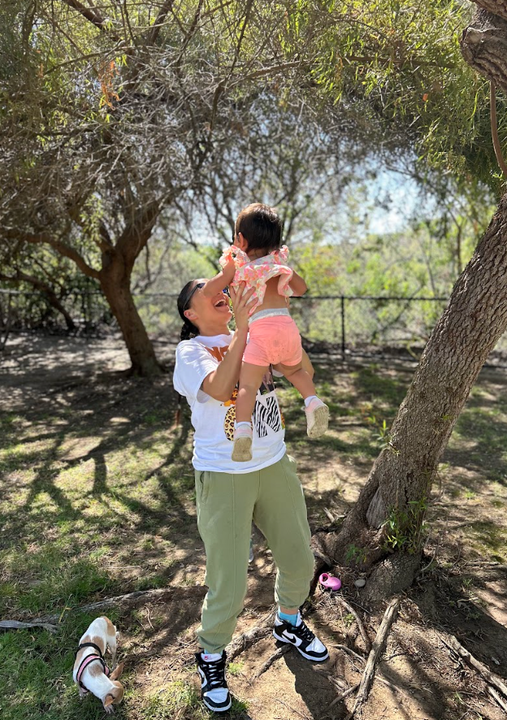 Adult lifting a child outdoors under trees in a sunny park, with a small dog nearby.