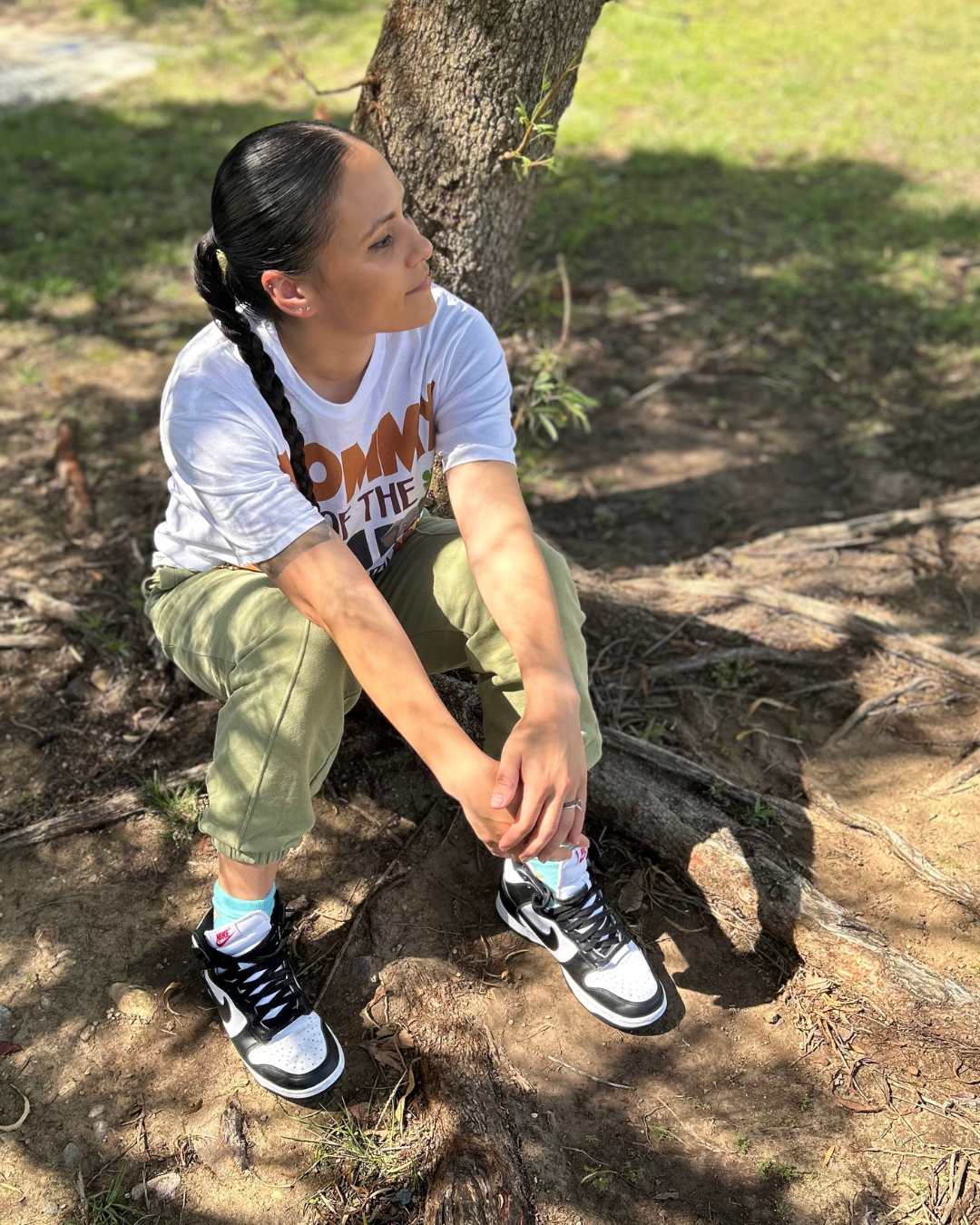 Person sitting on tree roots outdoors, wearing a white T-shirt, green pants, and black sneakers.