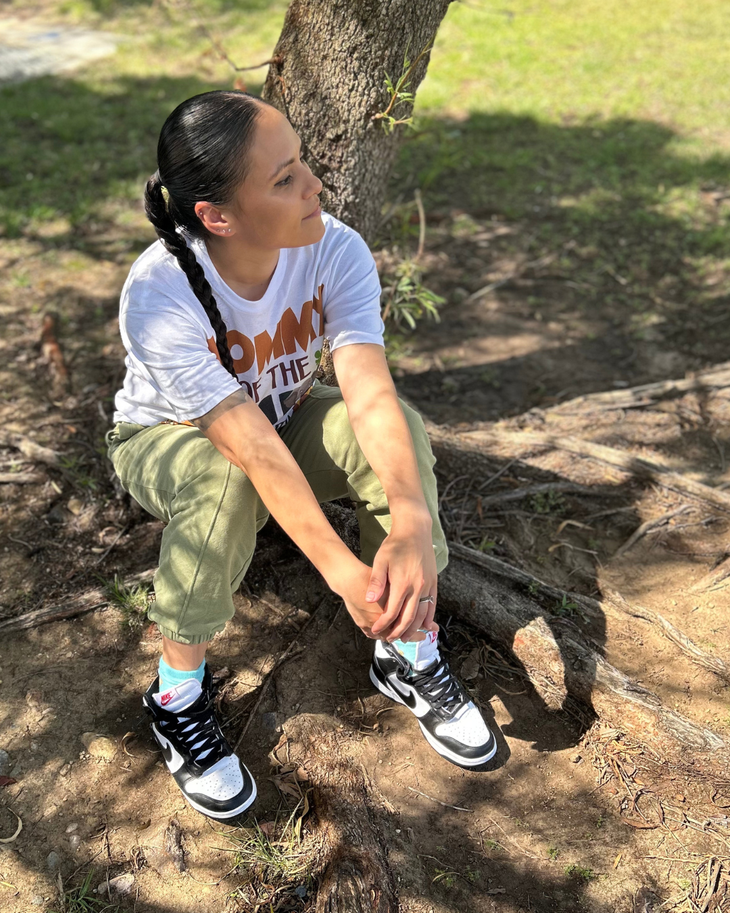Person sitting under a tree on a dirt path, wearing a white T-shirt, green pants, and black sneakers.