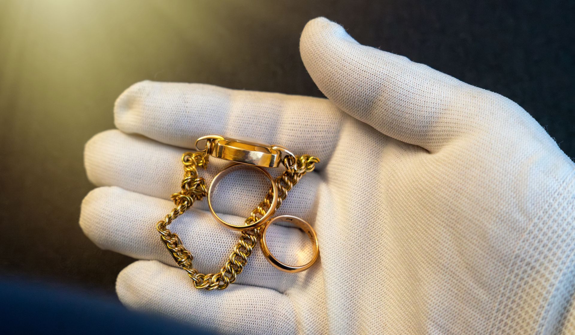 Gloved hand holding gold rings and a chain bracelet for jewelry appraisal.