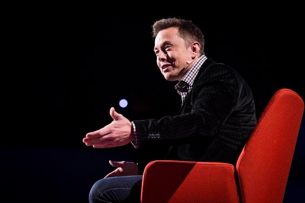 Elon musk is sitting in a red chair giving a speech.