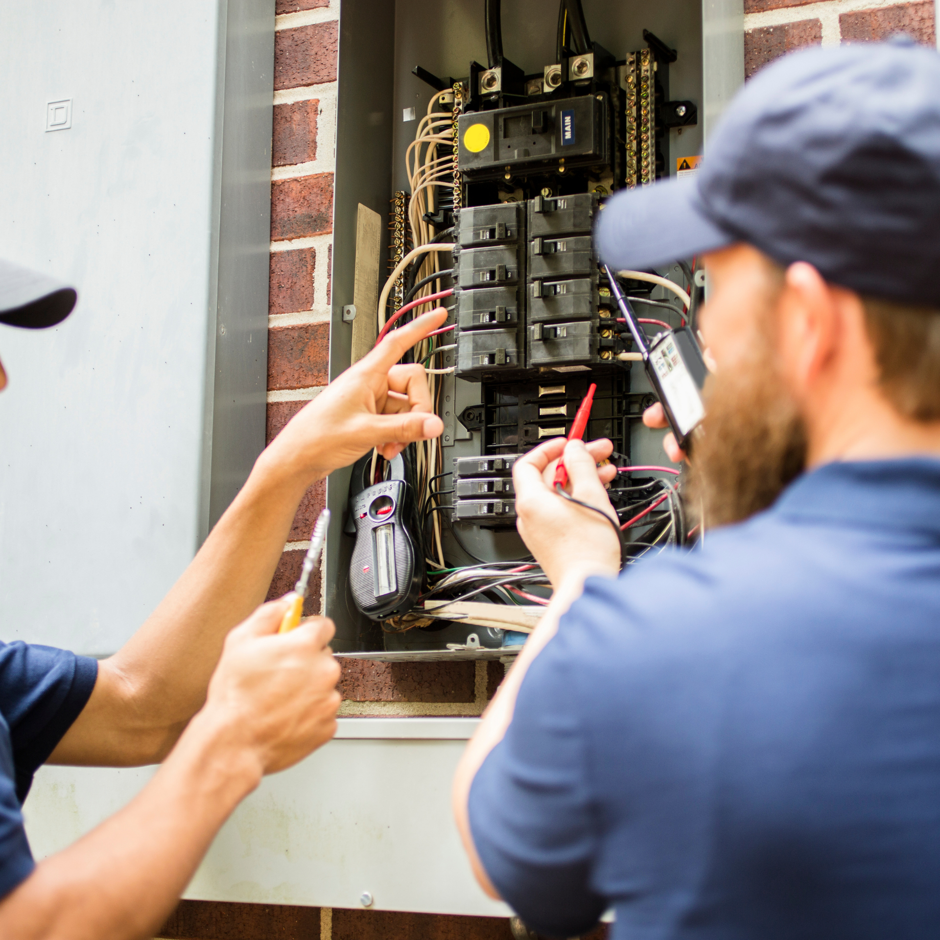 Two electricians working on an open electrical panel. One points, other uses a multimeter.