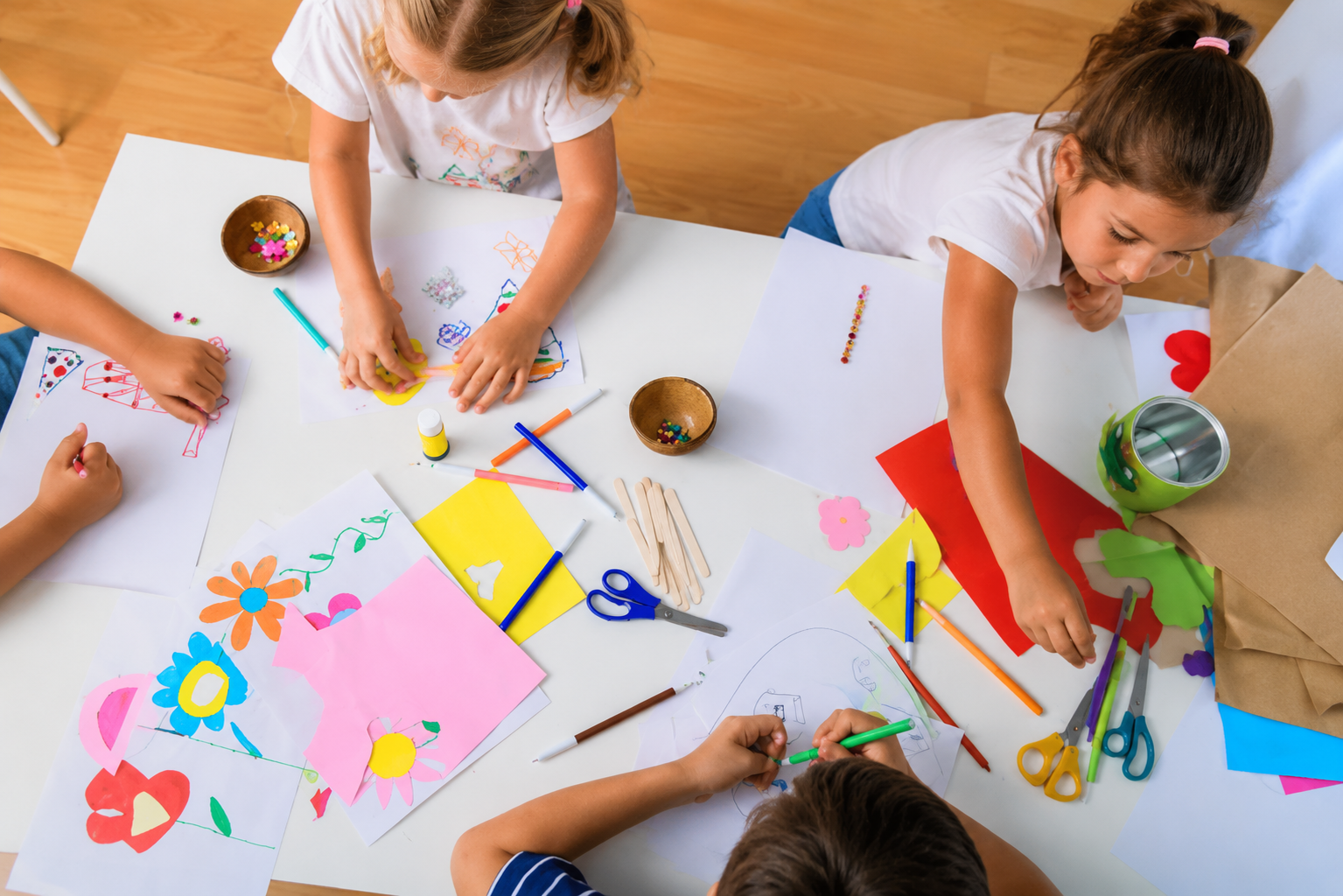 Des enfants peignent sur une grande bâche blanche, entourés de matériel de dessin coloré, sur un sol en bois.
