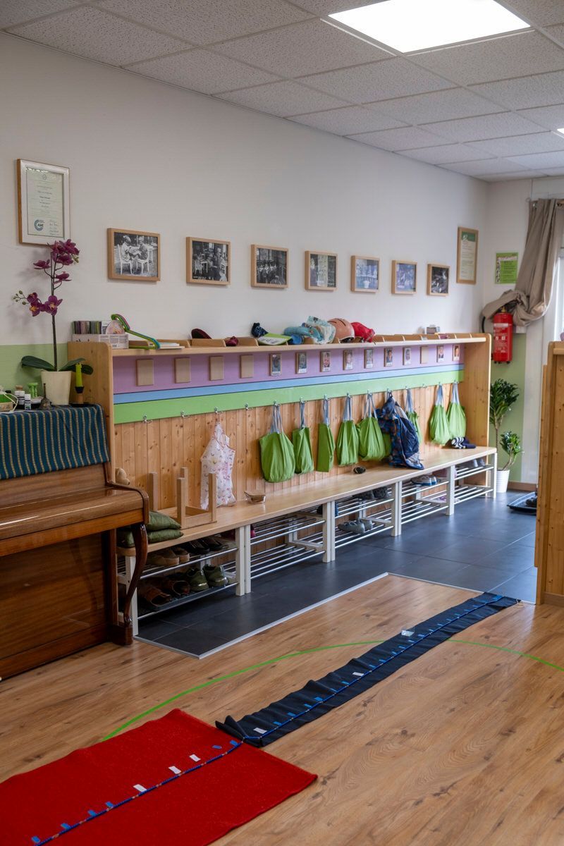 Couloir de classe avec casiers, sacs suspendus et tapis colorés sur un sol en bois