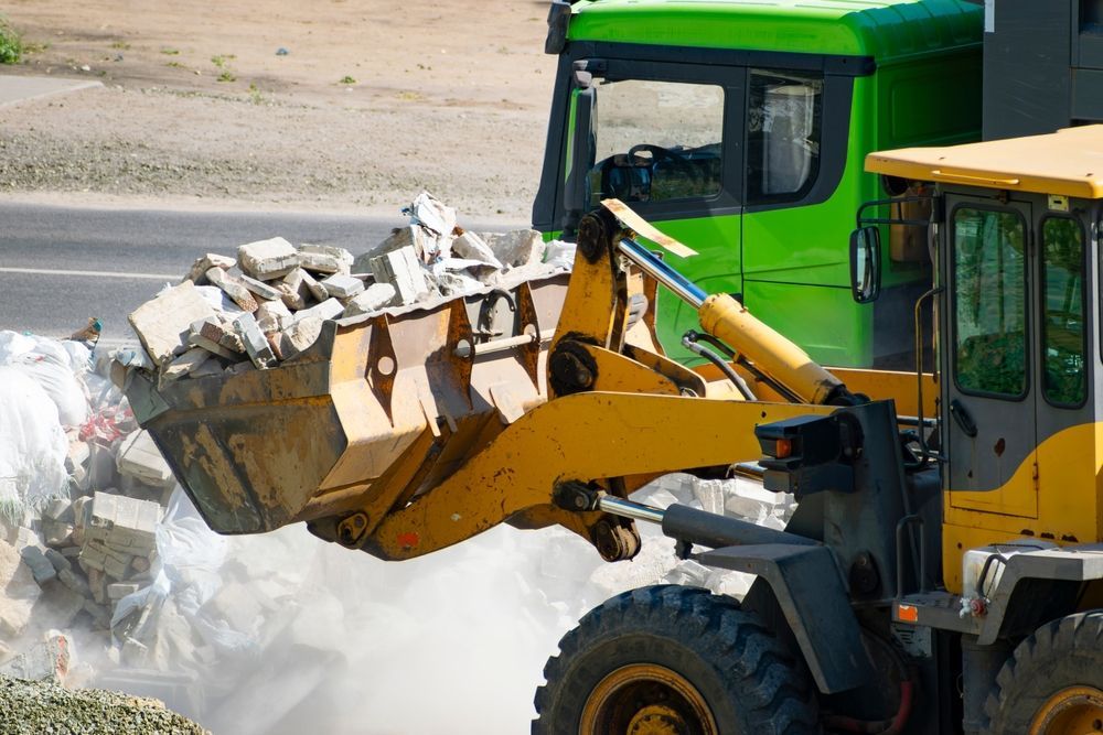 Yellow front loader dumping rubble near a green truck on a dusty construction site
