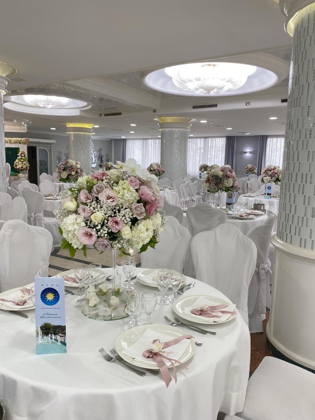 Elegante sala per banchetti con tavoli rotondi, tovaglie bianche, centrotavola floreali e sedie trasparenti.