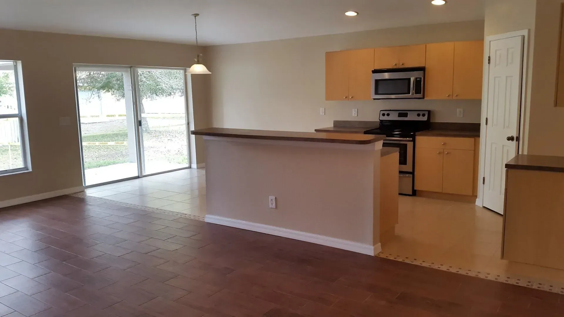 Interior of a home with open living space, kitchen, and sliding glass door. Hardwood floors, tan walls, and wood cabinets.
