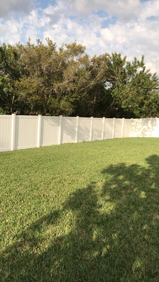 White vinyl fence in a grassy yard, trees in the background, blue sky with clouds.