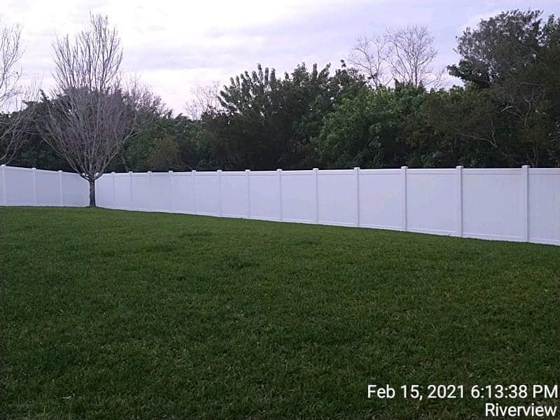 White fence in a grassy backyard with trees in the background. Cloudy sky.