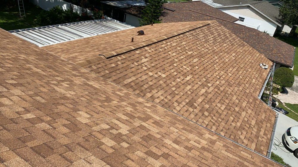 Brown asphalt shingle roof on a house, with a glimpse of the surrounding neighborhood and a car parked in front.