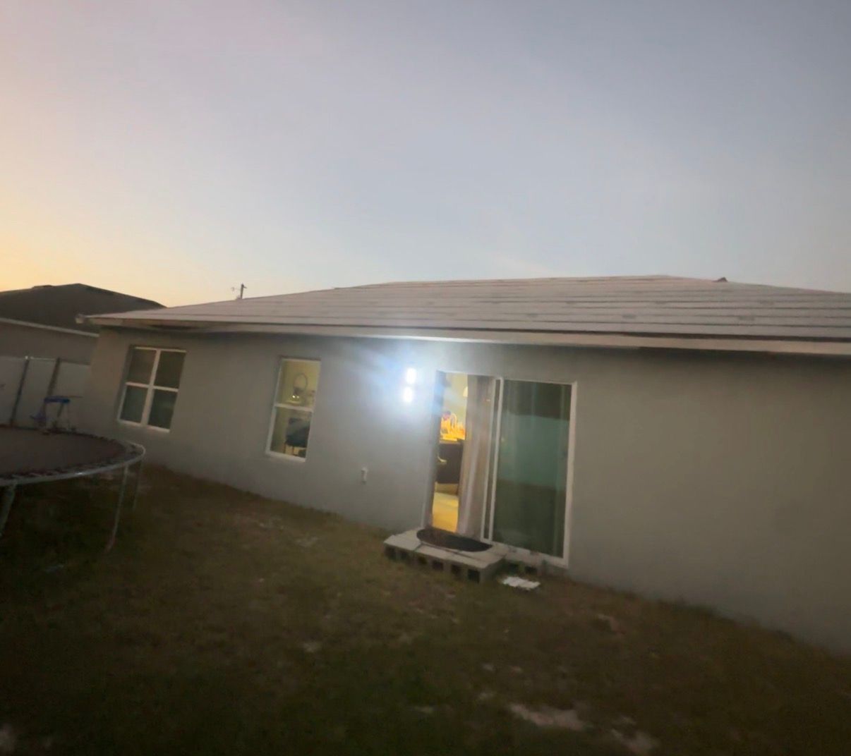 Back of a house at dusk. Gray siding, solar panels, sliding glass door, trampoline, and sparse grass.