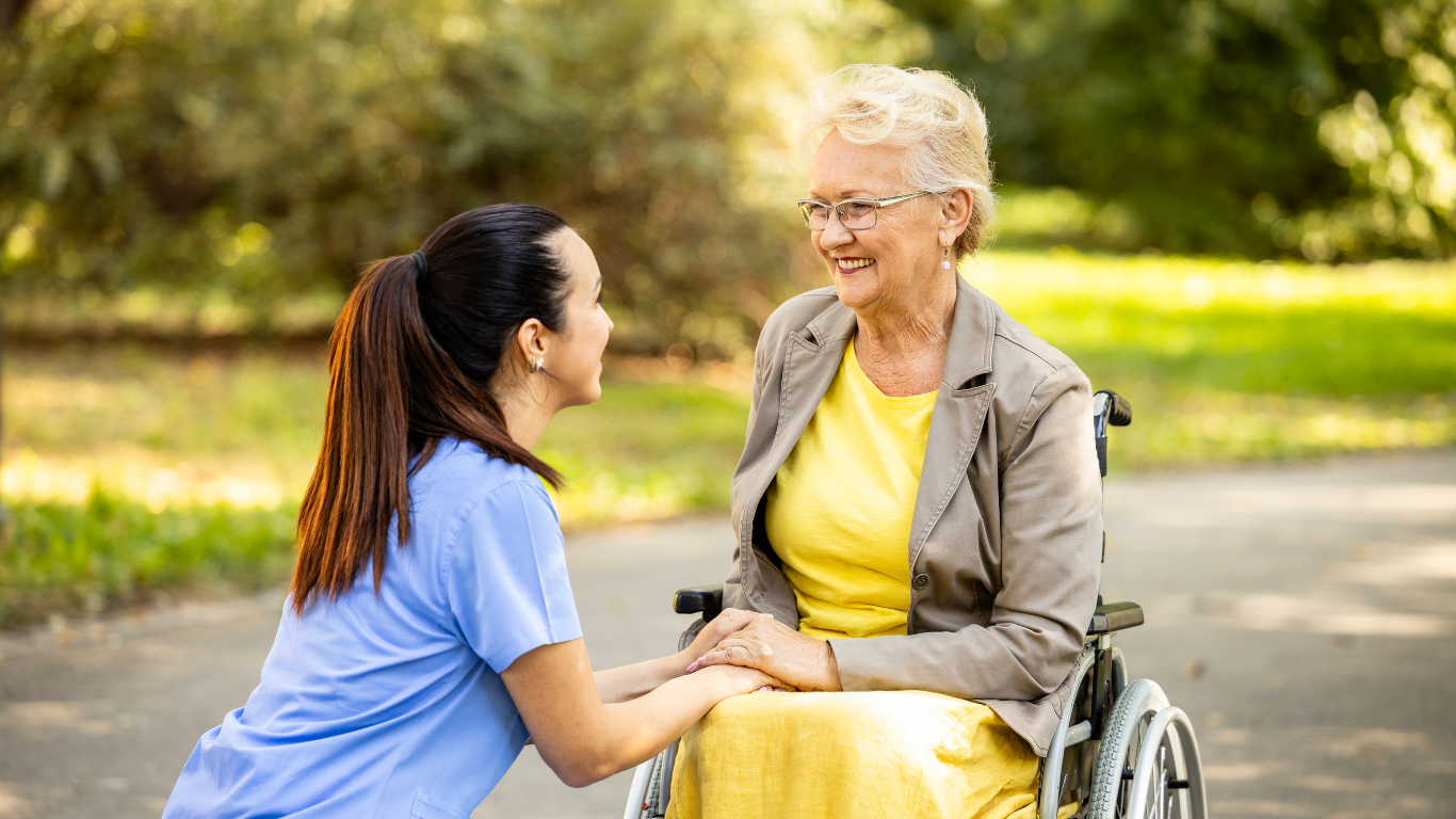 Caregiver kneels beside a wheelchair user in a bright room, adjusting the person’s shoe.
