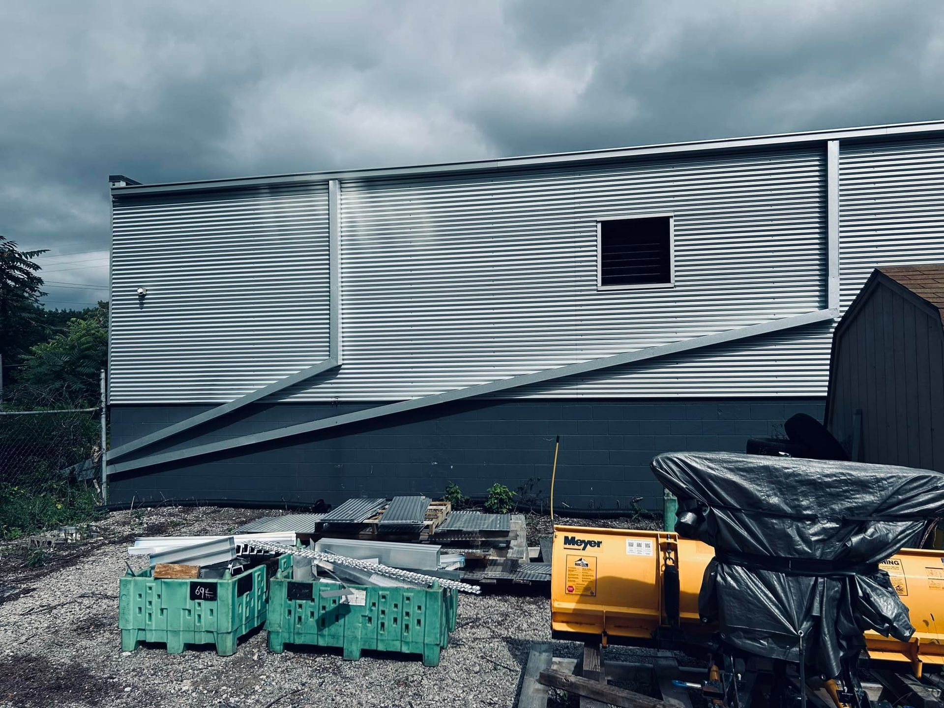 Damaged corrugated metal building with a bent wall, debris, and green crates in front under a cloudy sky